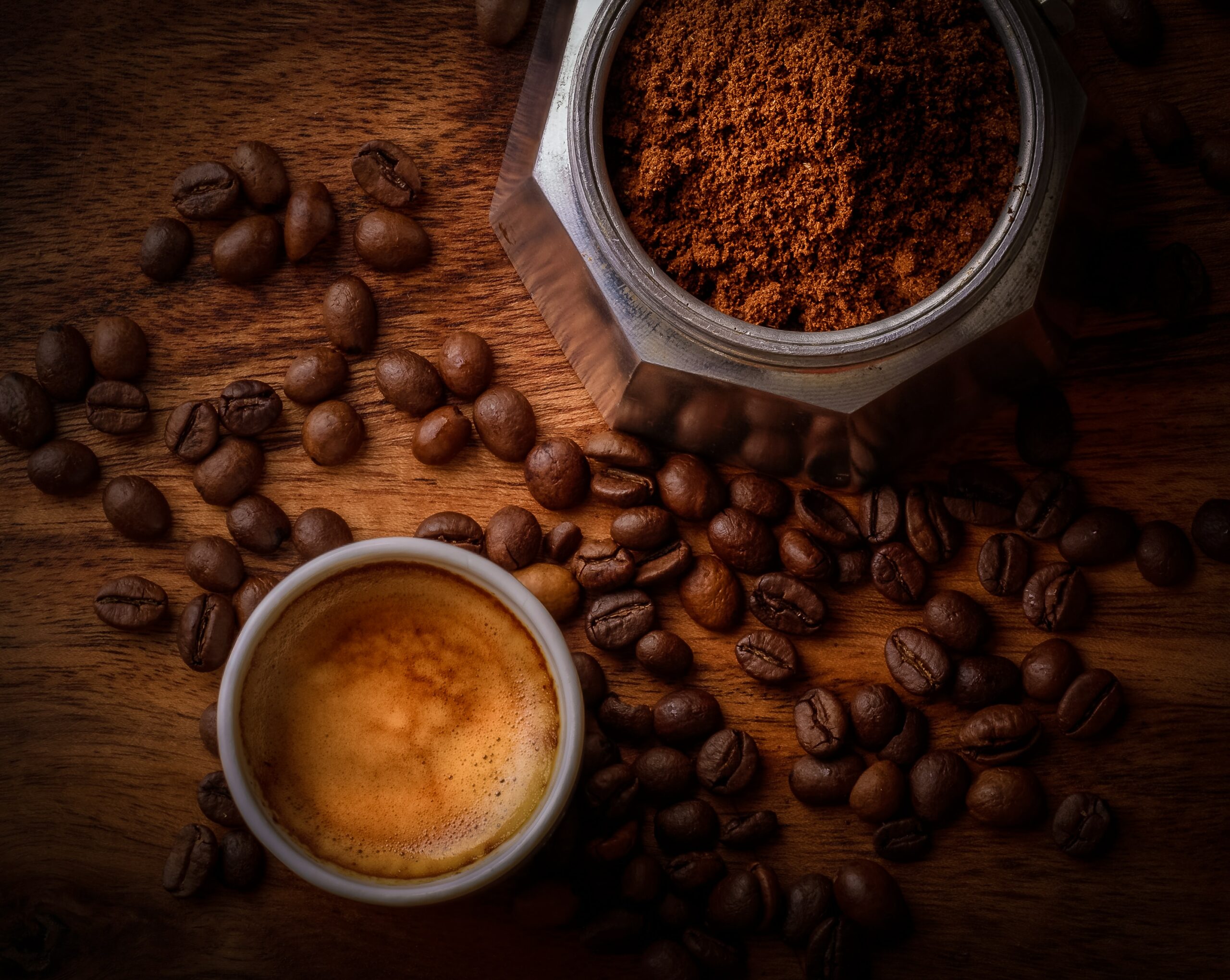 Kaffeebohnen auf einem braunen Holztisch. In der unteren Bildhälfte eine Tasse Kaffee, in der oberen Bildhälfte Kaffeesatz in einem Espressokocher.